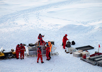 expédition scientifique de 7 hommes en rouge et un homme en orange inspectant des grosses caisses de matériels