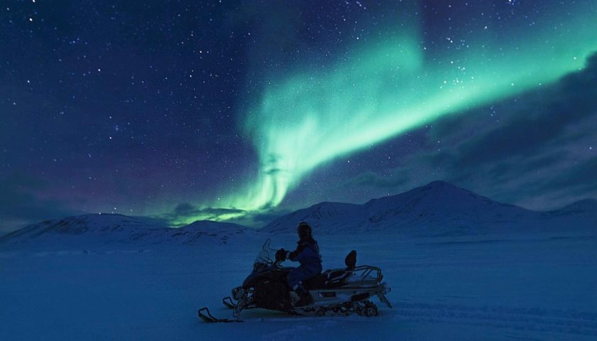 personne en moto-neige admirant une aurore boréale verte au dessus des montagnes